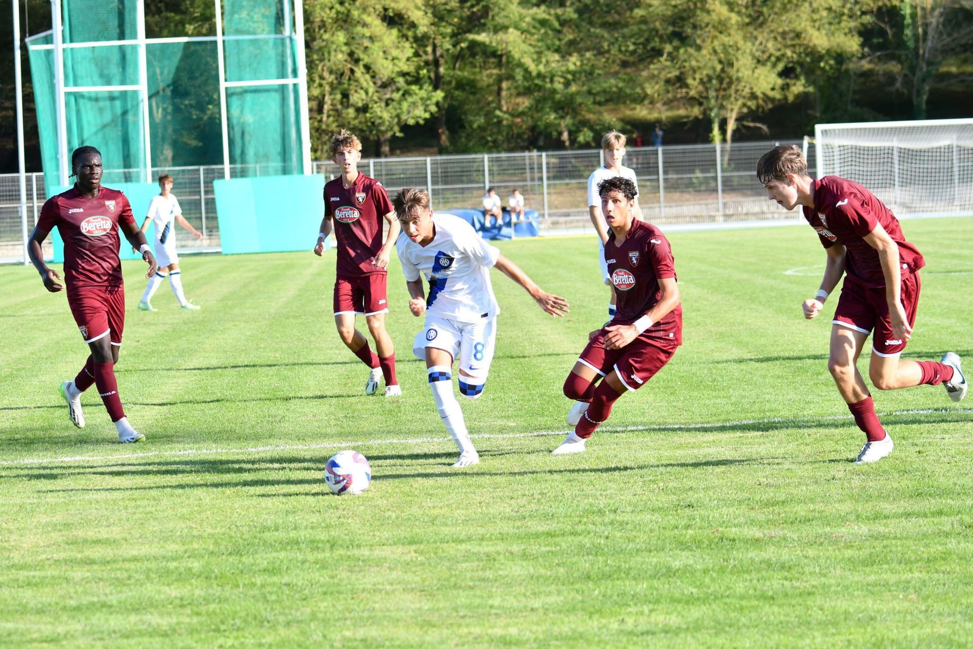 Giocatori di calcio con magliette bordeaux e bianche giocano su un campo verde.