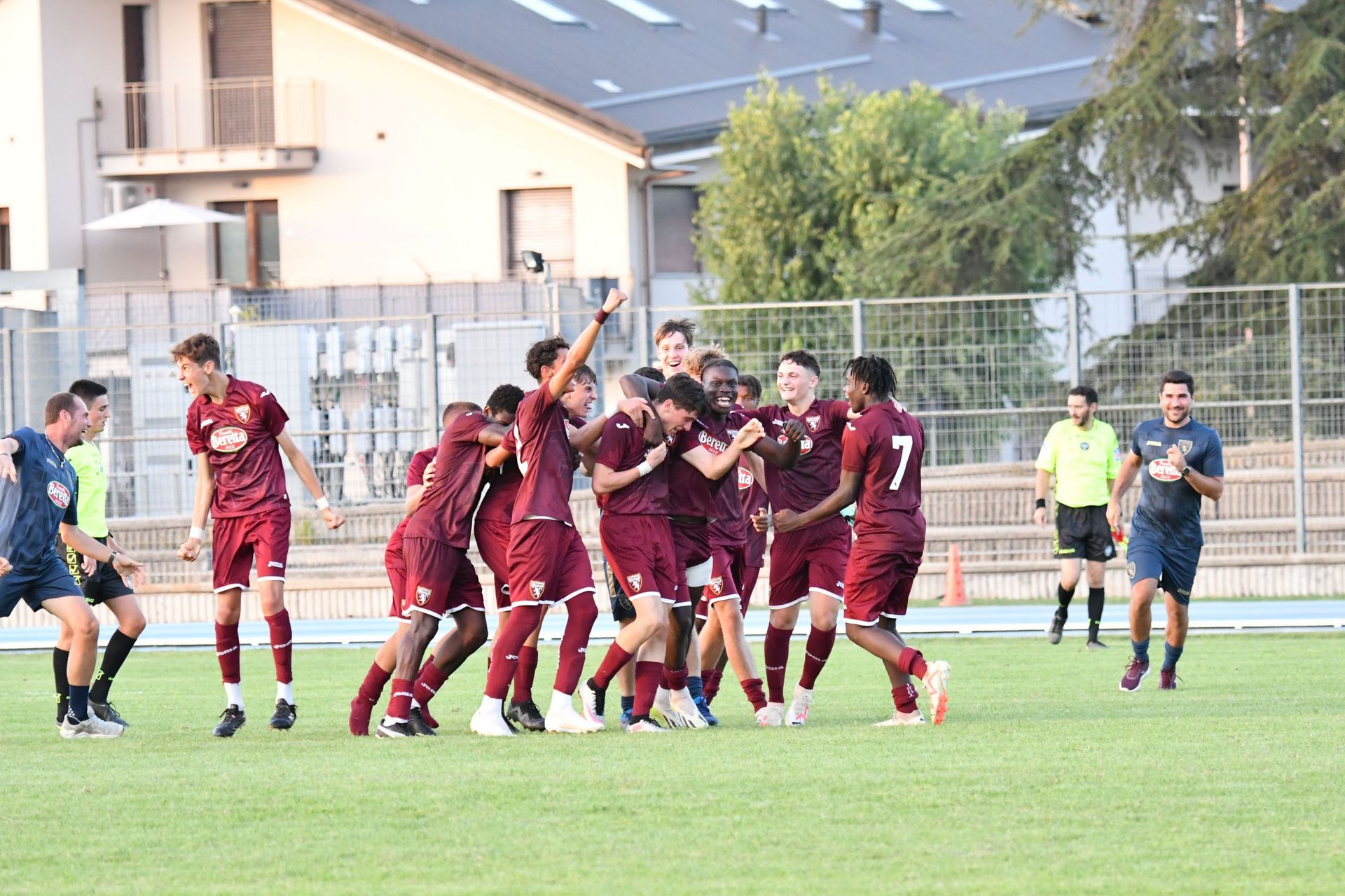 La squadra di calcio in maglia bordeaux festeggia su un campo verde. I giocatori si stringono, alcuni esultano. Gli arbitri osservano sullo sfondo.