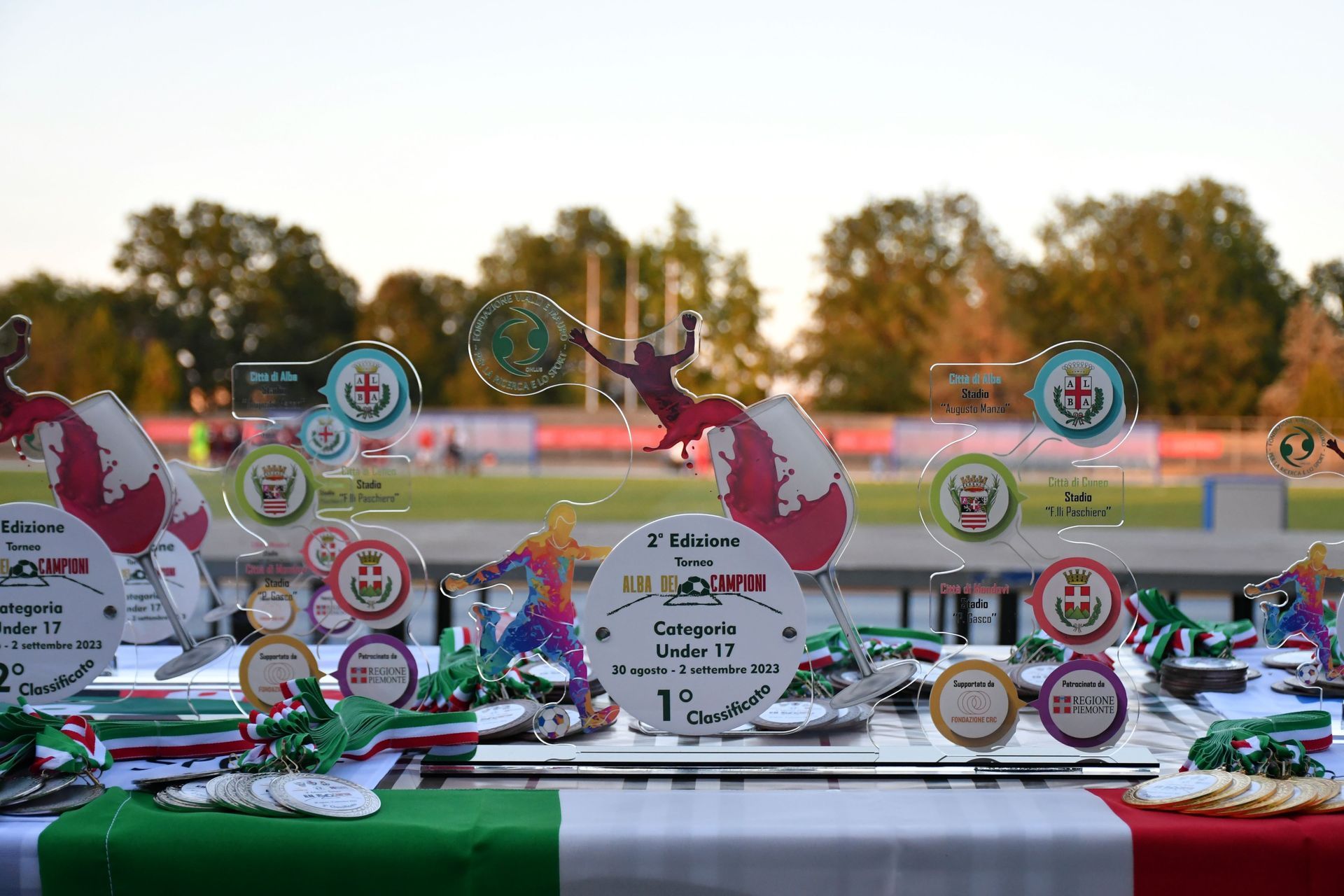 Trofei e premi su un tavolo ornato da una bandiera italiana, sullo sfondo di un campo sportivo.