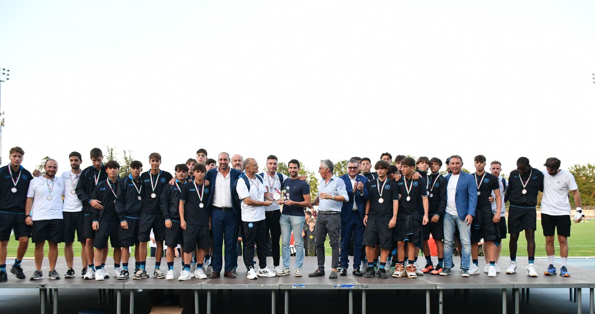 Squadra di calcio e dirigenti su un palco, alcuni con i trofei in mano, all'aperto. La squadra indossa divise nere, altri abiti formali.