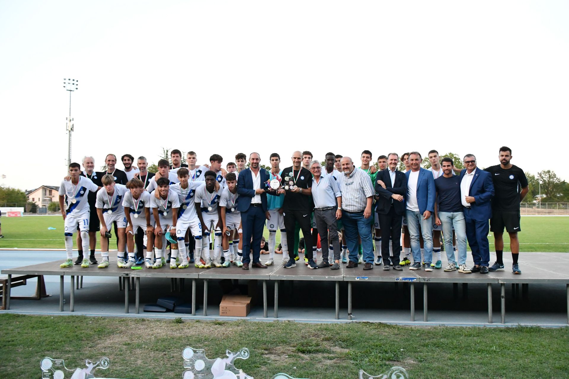 Squadra di calcio e dirigenti su un palco, in posa con i trofei. Ambientazione esterna, luce del giorno.