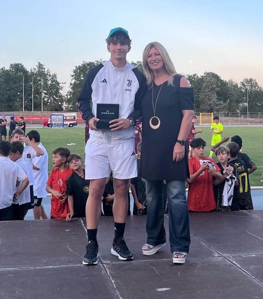 Un giovane uomo con giacca sportiva e una donna posano sul palco; sullo sfondo un campo da calcio.