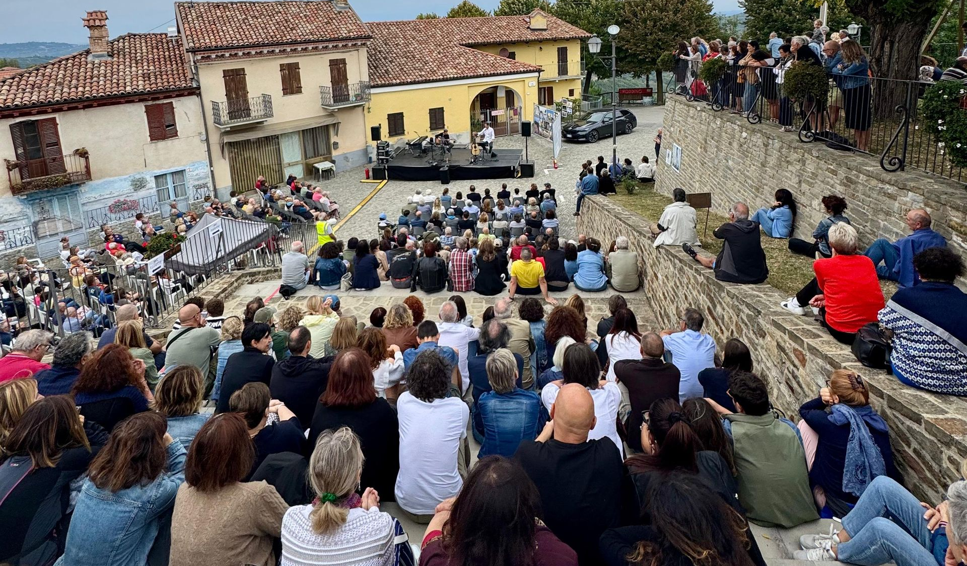 Persone sedute all'aperto assistono a un evento su un palco nella piazza di un villaggio europeo.