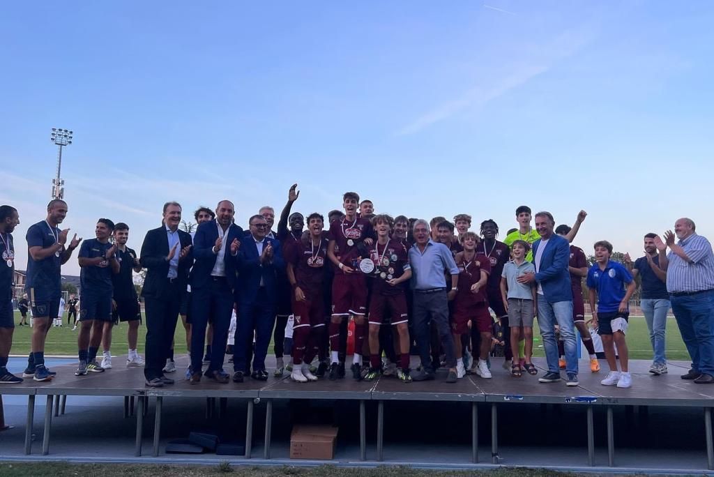 La squadra di calcio festeggia su un palco all'aperto sotto un cielo azzurro, tenendo in mano i trofei e posando per una foto di gruppo.
