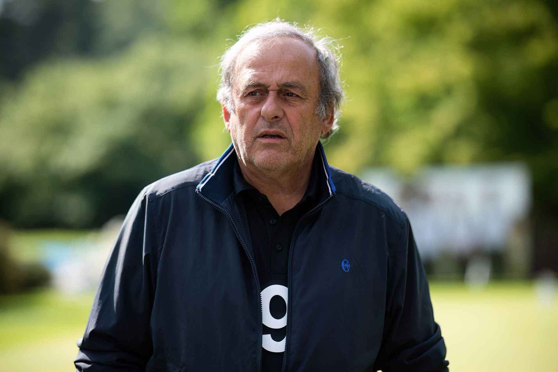 Uomo con giacca blu e camicia nera, guarda di lato, all'esterno, con alberi sullo sfondo.