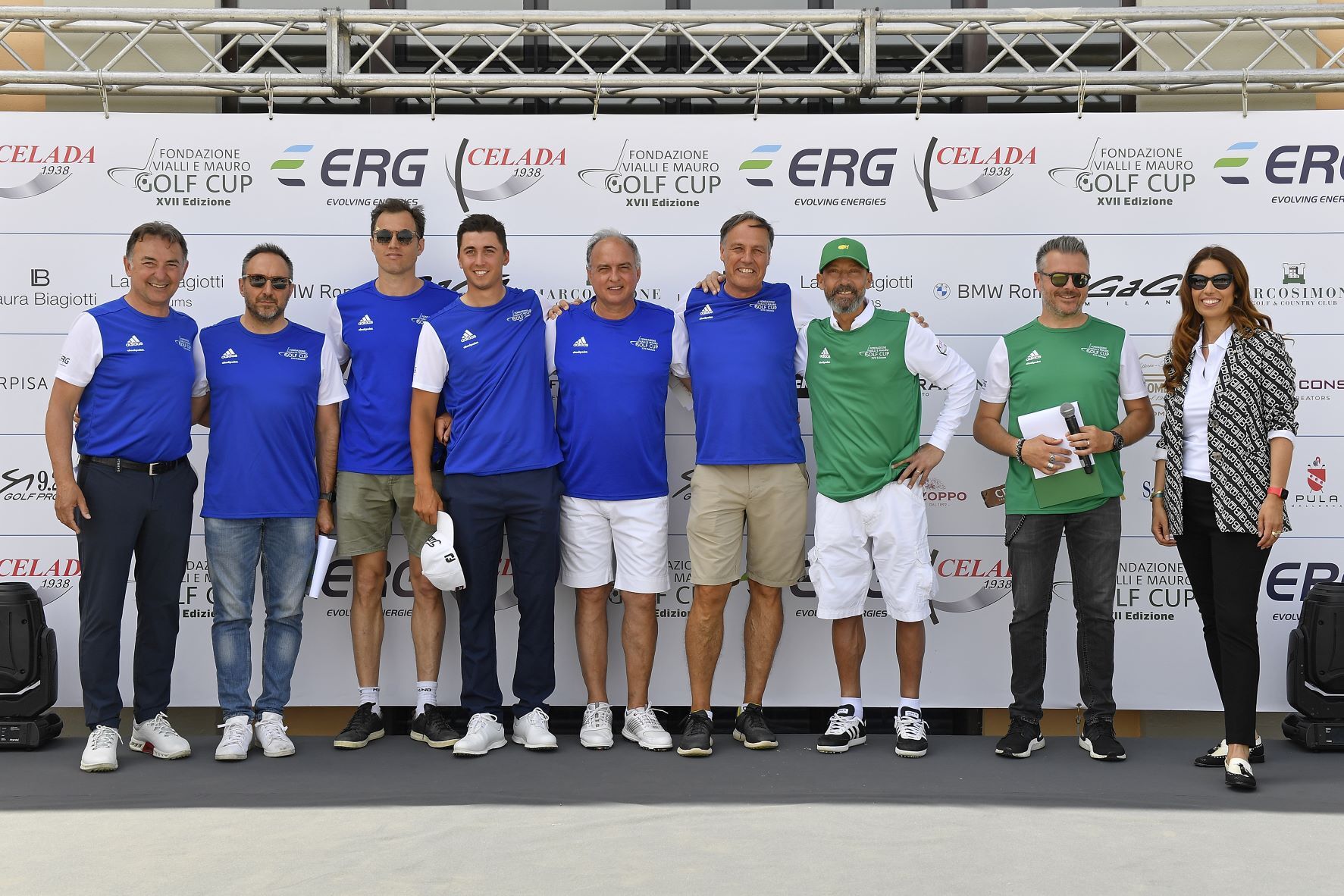 Gruppo di persone con magliette blu e verdi, in posa su un palco con uno striscione degli sponsor.