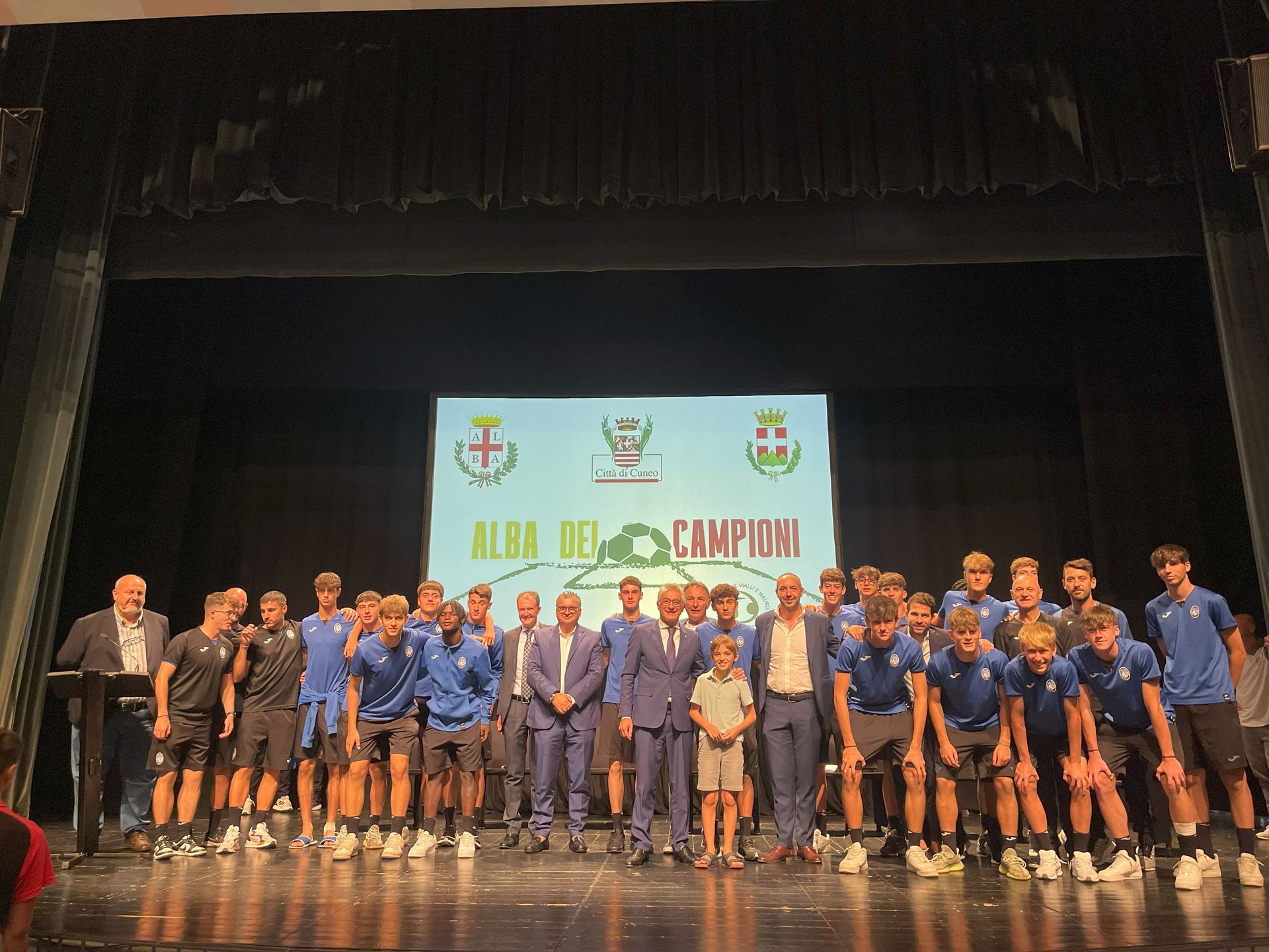 Gruppo di persone in posa su un palco, alcune in tenuta sportiva blu, con uno striscione dietro di loro che recita