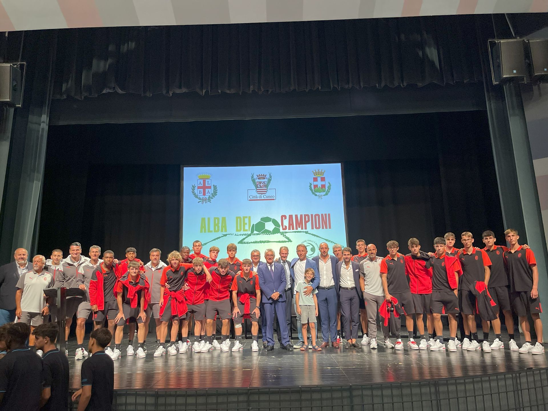 Foto di gruppo sul palco: una squadra di calcio in divisa rossa e nera posa con i dignitari davanti a uno striscione.