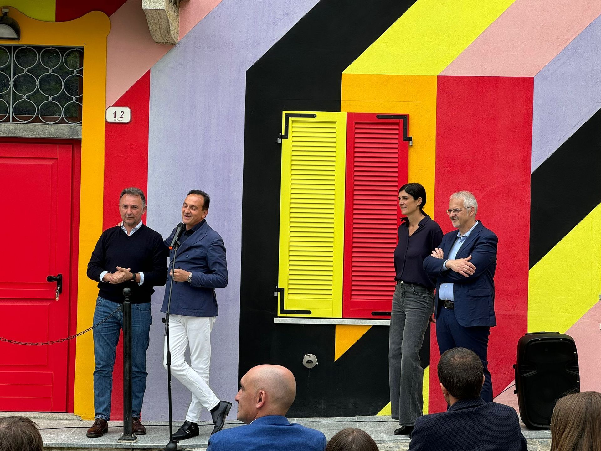 Quattro persone sono in piedi su un palco di fronte a un edificio colorato. Una di loro parla al microfono.