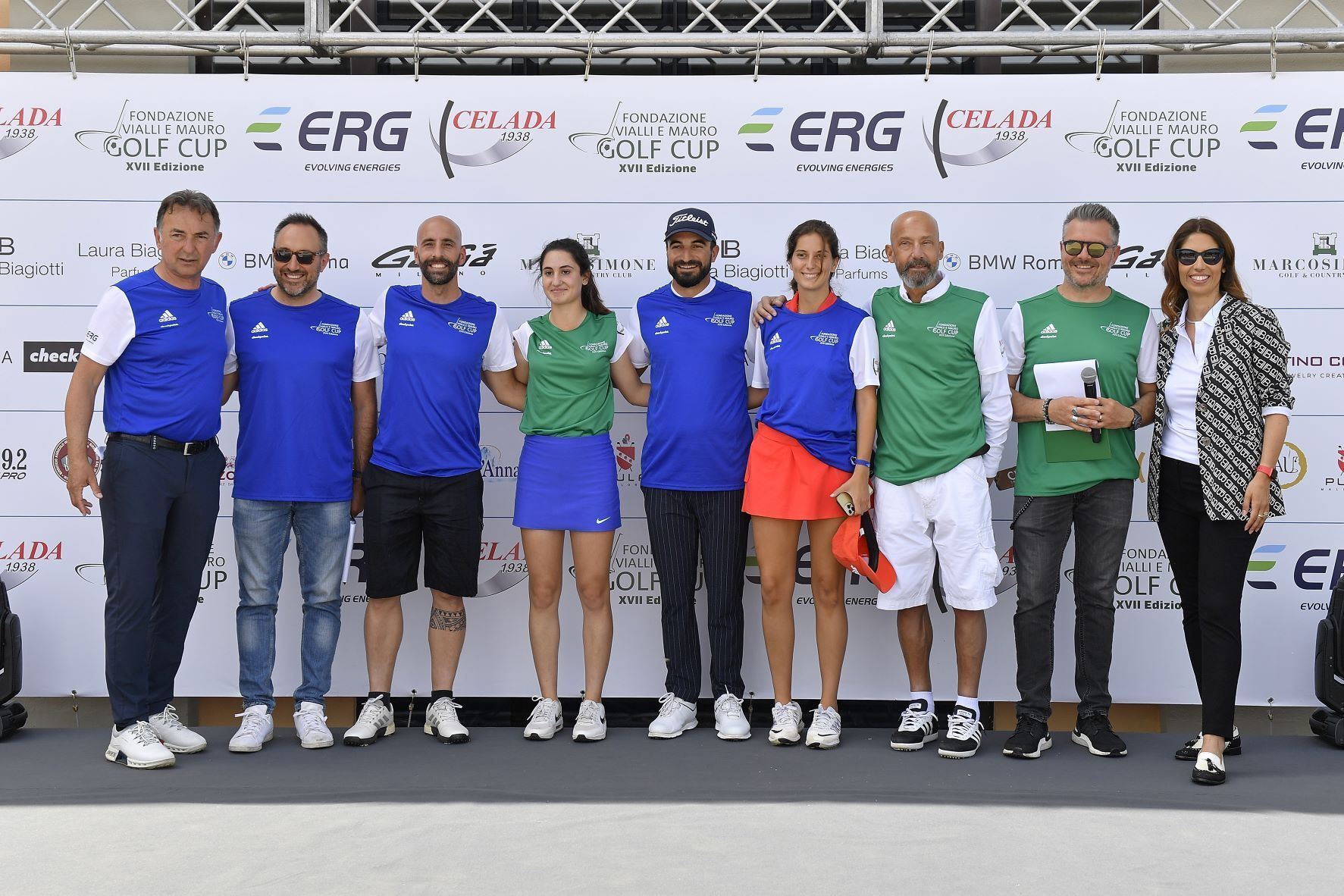 Gruppo di persone in piedi su un palco durante un evento di golf, alcune delle quali indossano magliette e pantaloncini blu o verdi.