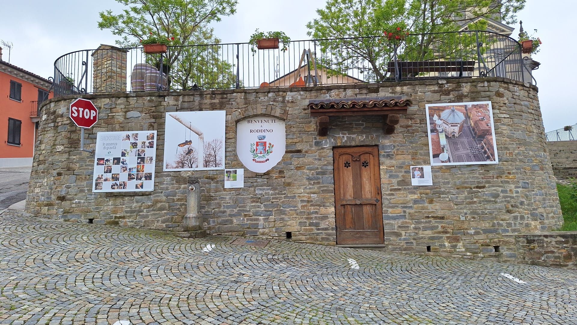 Edificio in pietra con manifesti, una porta in legno e una terrazza con piante, posto su ciottoli.