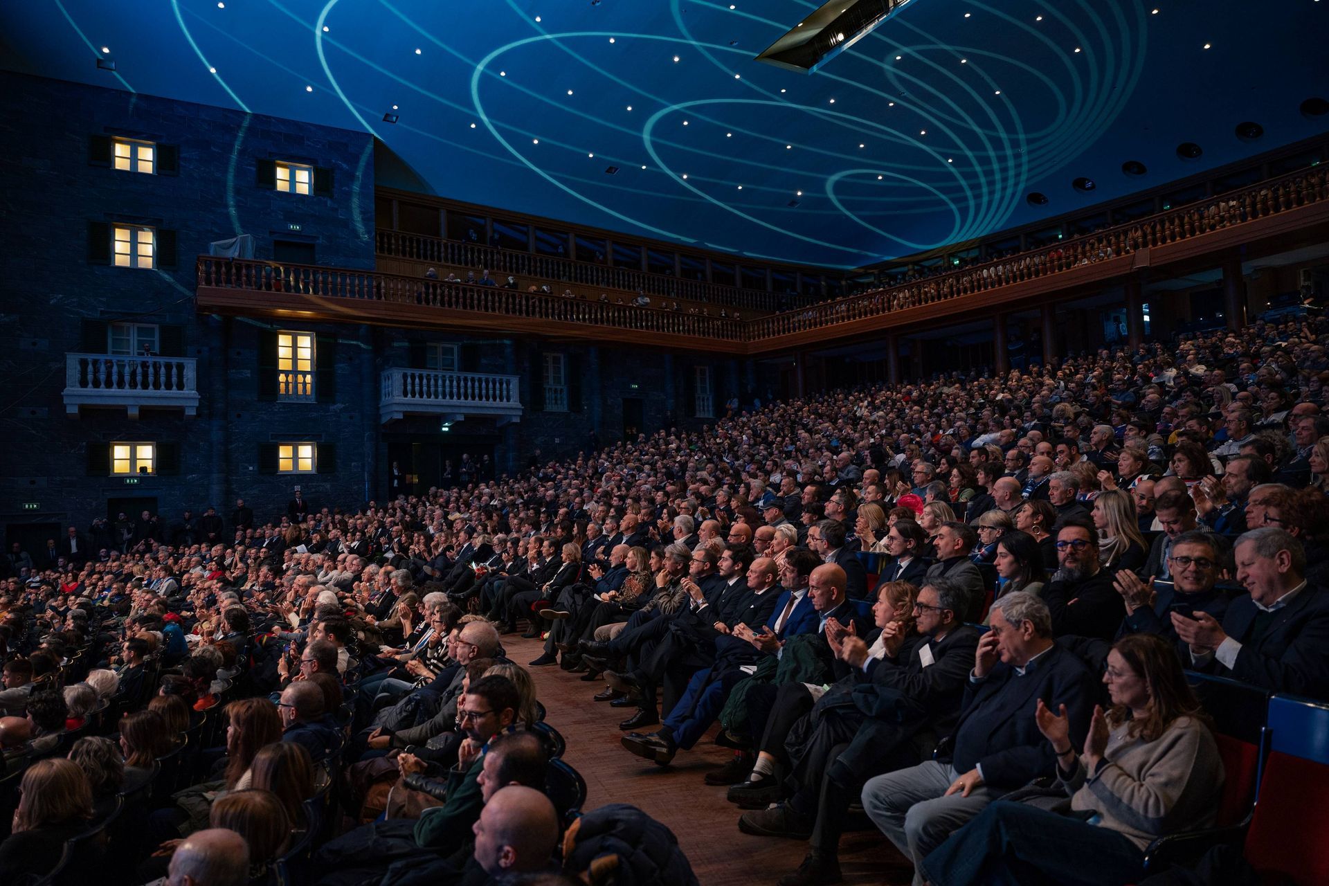 Pubblico in un grande teatro, con lo sguardo rivolto verso l'alto verso il soffitto blu, con luci e balconi.