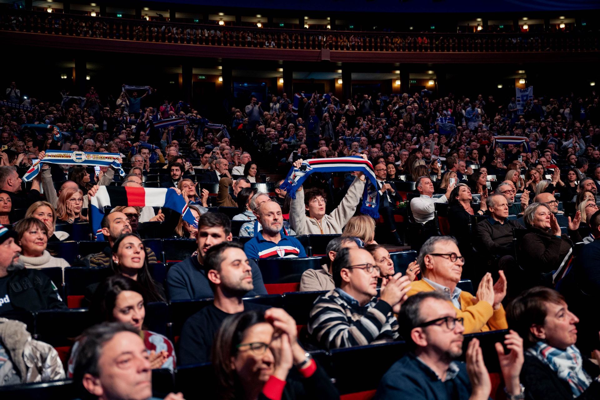 Una grande folla applaude in un'arena. Molti tengono sciarpe blu, bianche e rosse.