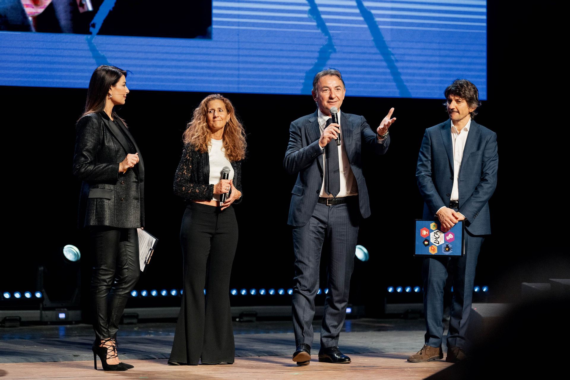 Quattro persone su un palco con uno schermo alle spalle. Un uomo parla al microfono.