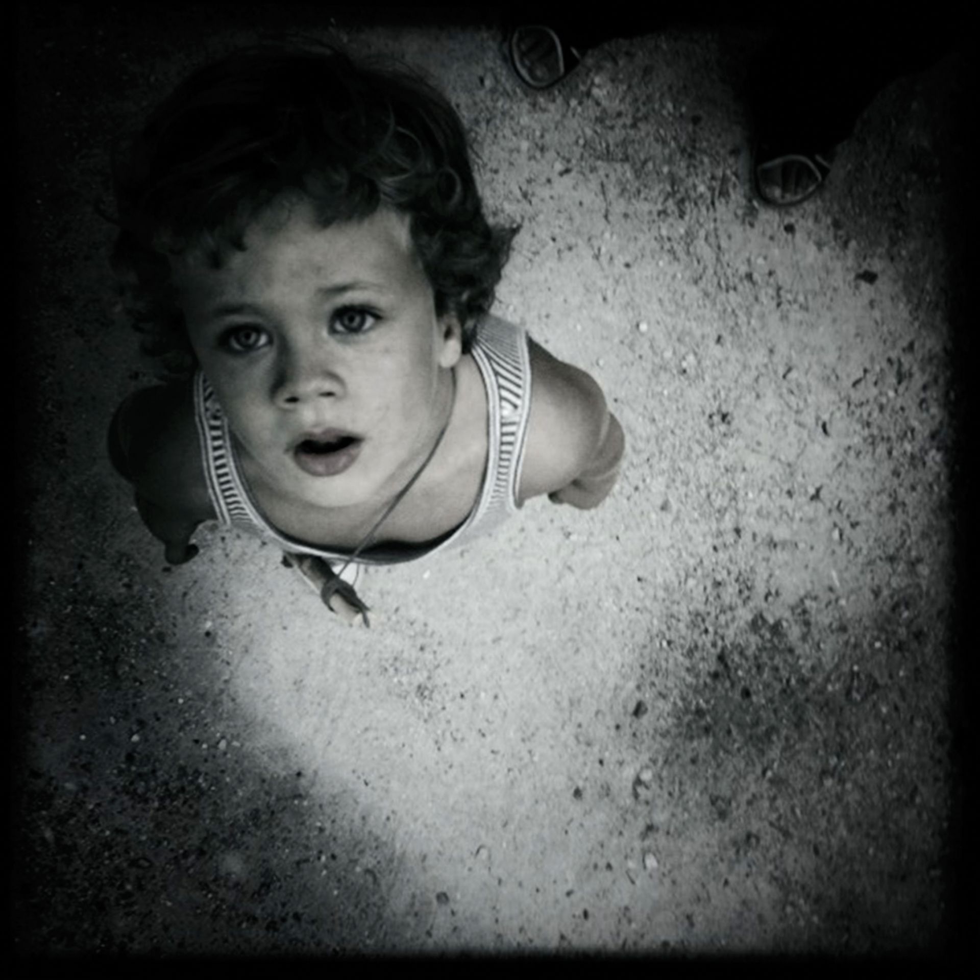 Bambino che guarda verso l'alto, a bocca aperta, su uno sfondo di cemento strutturato; fotografia in bianco e nero.