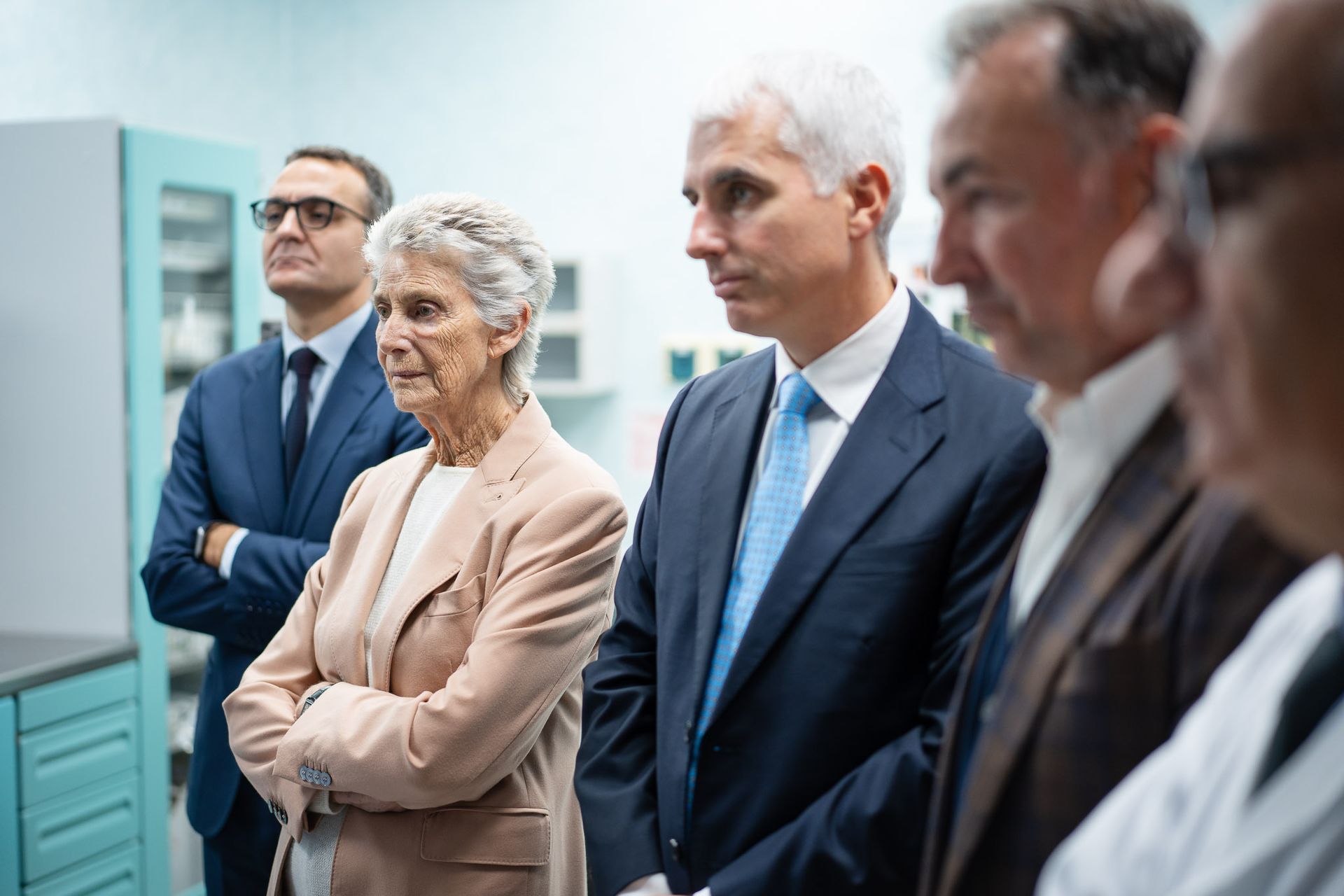 Persone in abiti da lavoro guardano intensamente qualcosa fuori dall'inquadratura; sullo sfondo, armadietti azzurri.