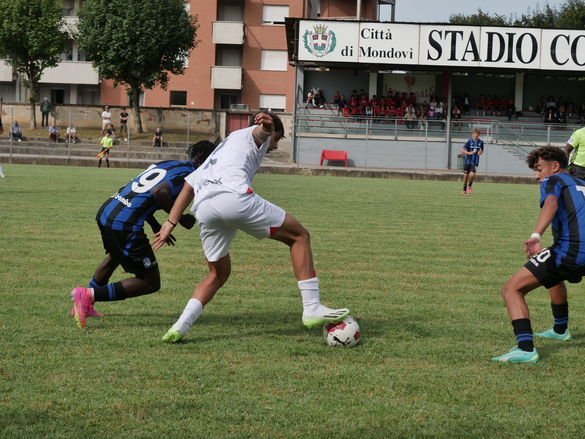 Giocatori di calcio su un campo erboso; uno in bianco supera in palleggio due in blu. Spettatori sullo sfondo.