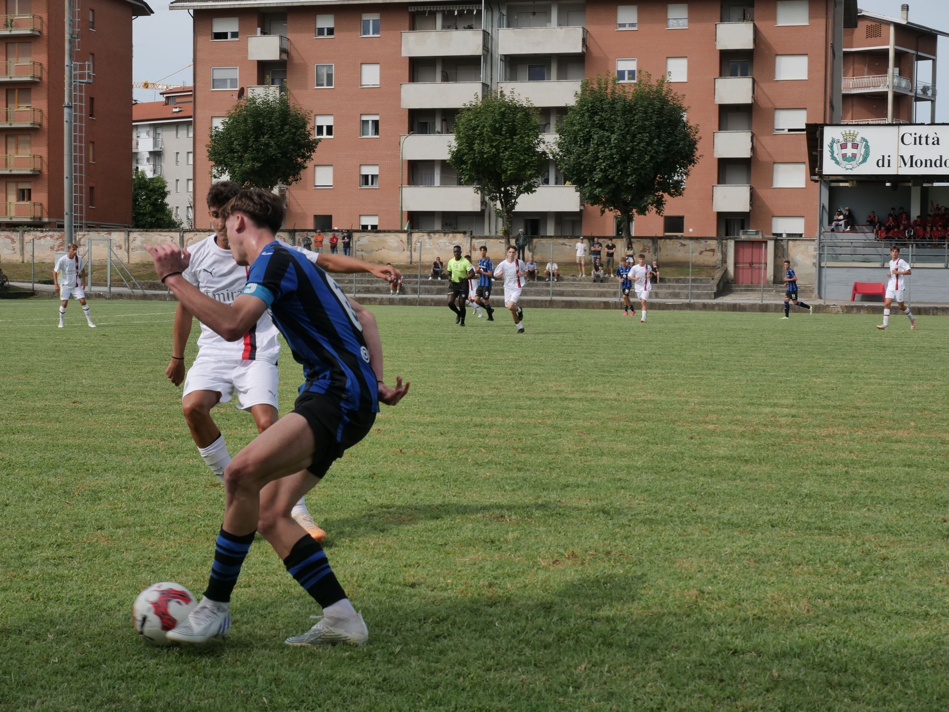Giocatori di calcio su un campo; uno in blu, nero e bianco calcia un pallone mentre un altro in bianco difende. Edifici sullo sfondo.