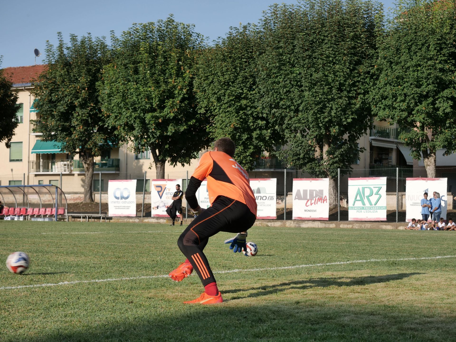 Portiere di calcio in uniforme arancione e nera che calcia la palla su un campo verde. Alberi e striscioni sullo sfondo.