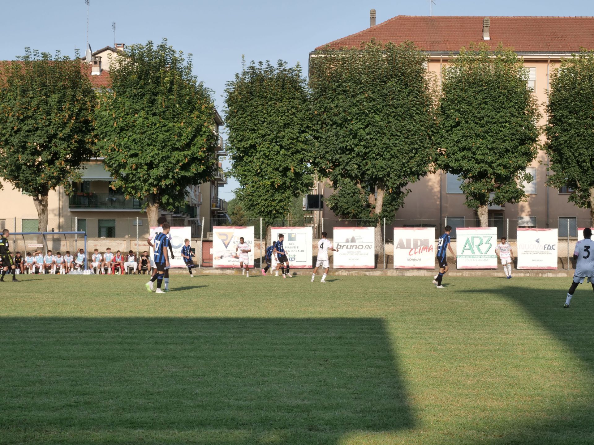 Partita di calcio su un campo verde con giocatori in blu e bianco. Edifici e spettatori sullo sfondo.
