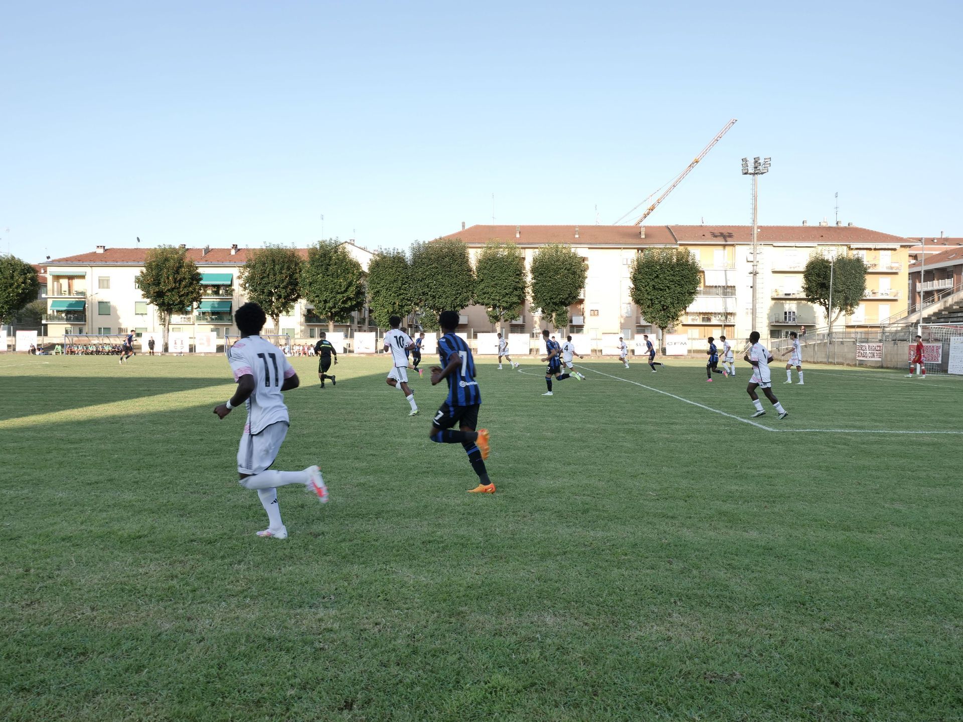 Giocatori di calcio su un campo verde con edifici sullo sfondo. I giocatori corrono.