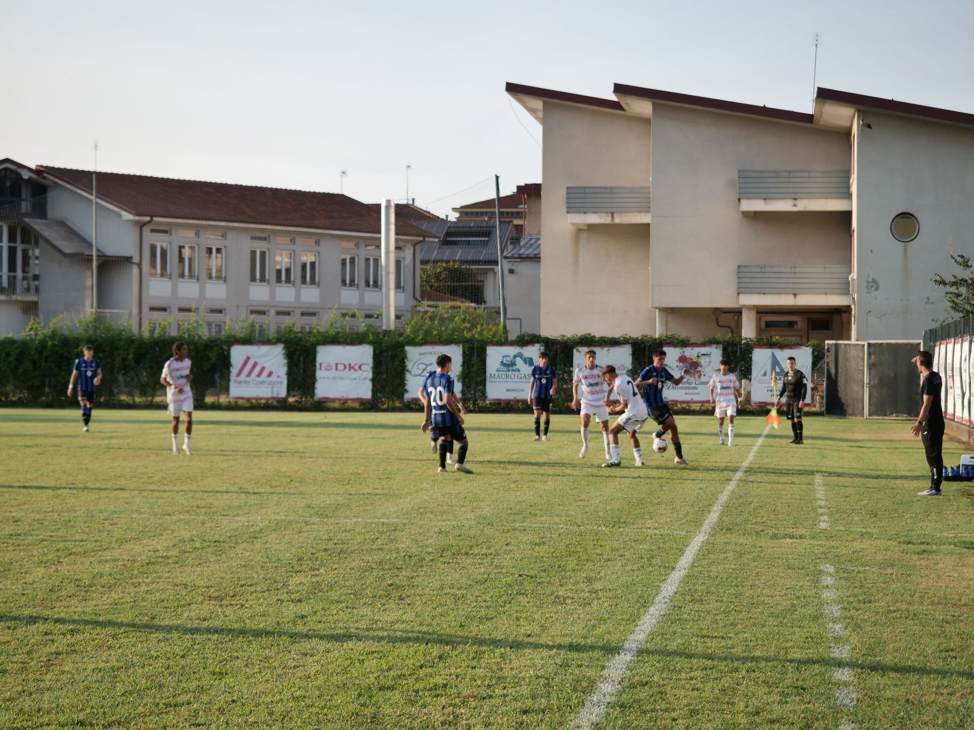 Partita di calcio in corso su un campo verde, giocatori in divisa bianca e blu vicino agli edifici.