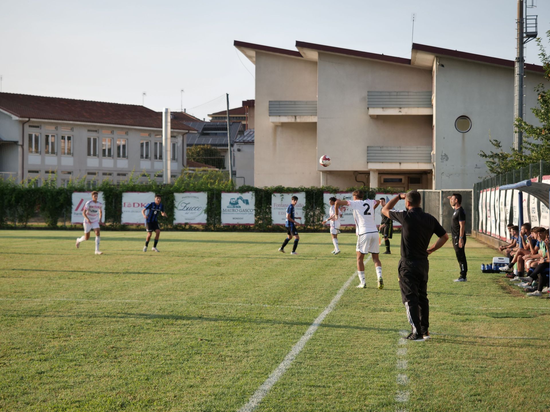 Partita di calcio in corso su un campo verde, giocatori in divisa bianca e blu, edifici sullo sfondo.