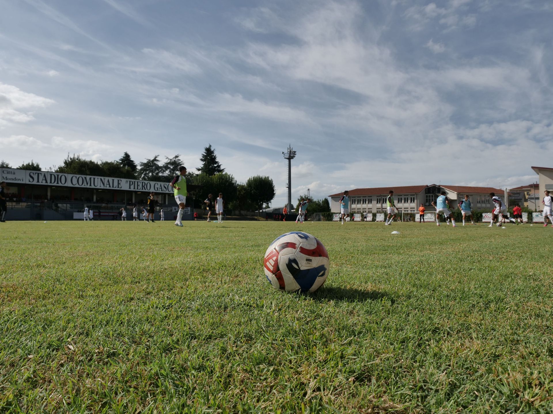 Pallone da calcio su un campo verde; i giocatori si allenano in lontananza sotto un cielo parzialmente nuvoloso.
