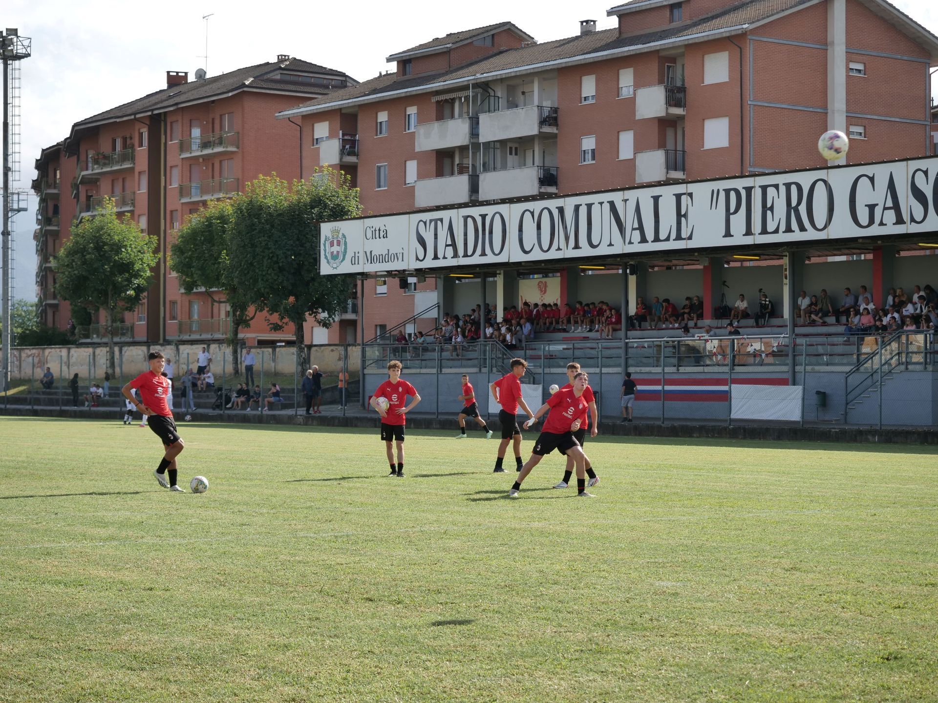 Giocatori di calcio con magliette rosse su un campo verde davanti a uno stadio con spettatori.