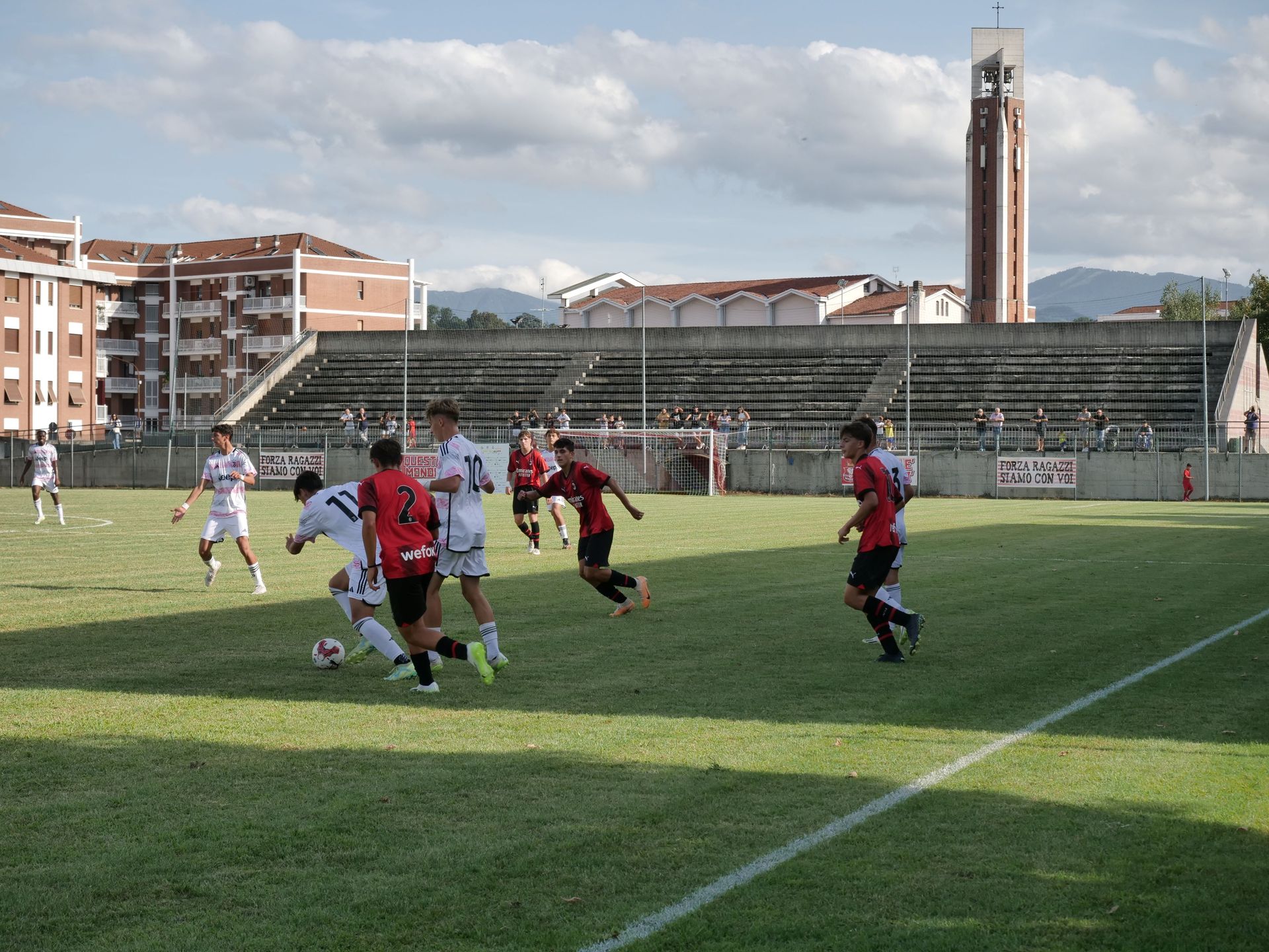 Partita di calcio su un campo verde, giocatori in divisa rossa e bianca. Spettatori sugli spalti. Sullo sfondo, l'alta torre dell'orologio.