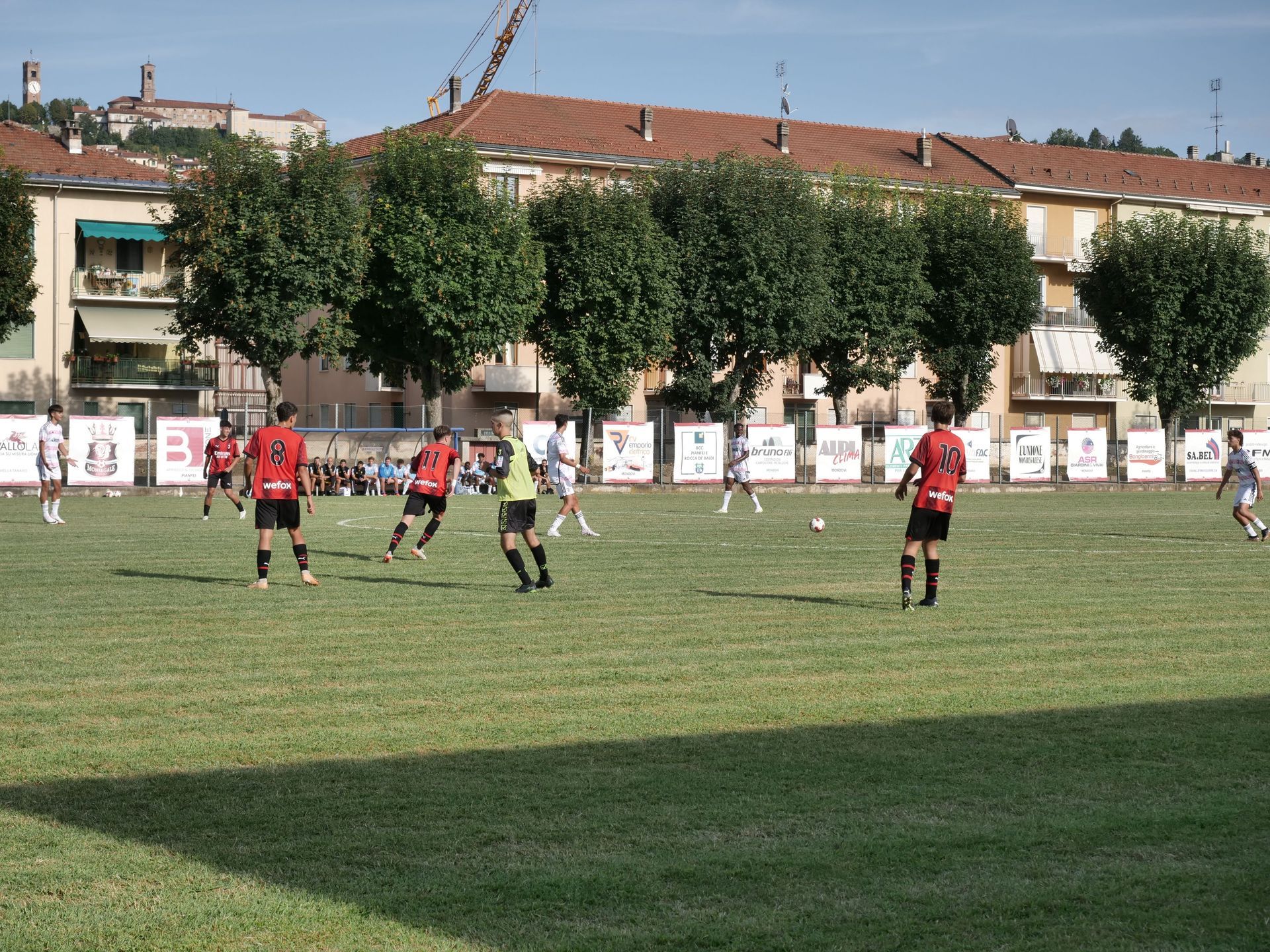 Giocatori di calcio con maglie rosse e bianche su un campo erboso, con edifici sullo sfondo.
