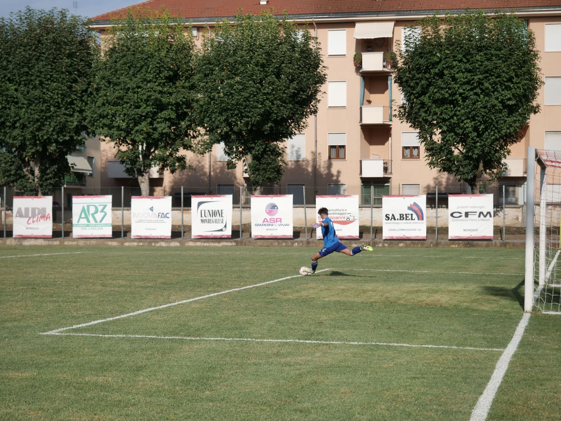 Calciatore in blu che calcia la palla su un campo verde verso una porta. Sullo sfondo ci sono striscioni.
