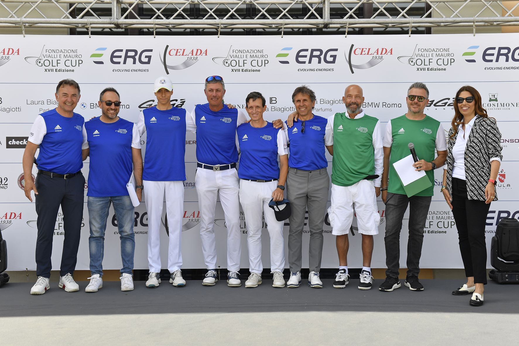 Gruppo di persone in posa su un palco, alcune con magliette blu e bianche. Dietro di loro si vedono i loghi degli sponsor.
