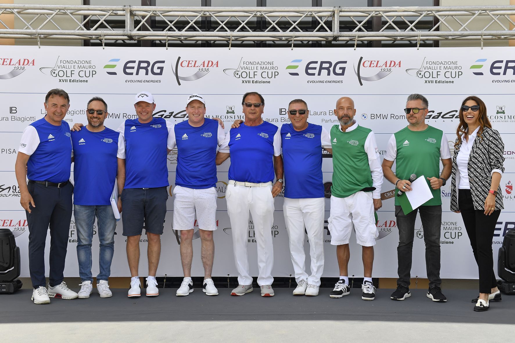 Gruppo di persone in posa su un palco davanti a uno striscione. Molti indossano magliette blu o verdi e pantaloni bianchi.