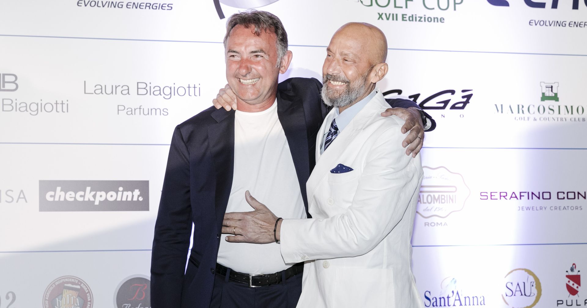 Due uomini in giacca e cravatta, sorridenti e abbracciati durante un evento all'aperto con i loghi degli sponsor.