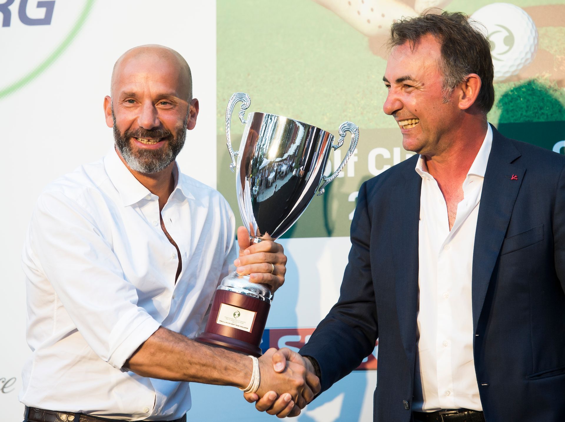 Uomo in camicia bianca che tiene in mano un trofeo e stringe la mano a un altro uomo in giacca, all'aperto.