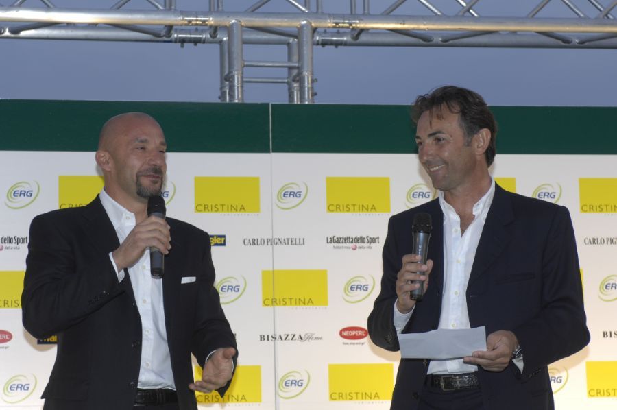 Due uomini in giacca e cravatta tengono in mano dei microfoni su un palco e parlano. Sullo sfondo ci sono i loghi aziendali.