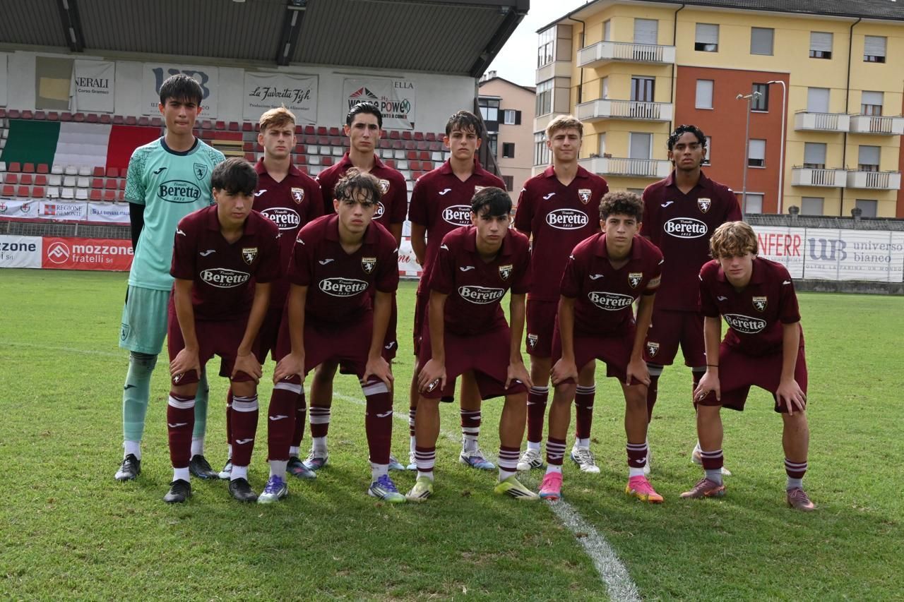 Quattro uomini sono in piedi su un palco, uno dei quali parla al microfono. Sono all'aperto, con un campo da calcio e alberi sullo sfondo.