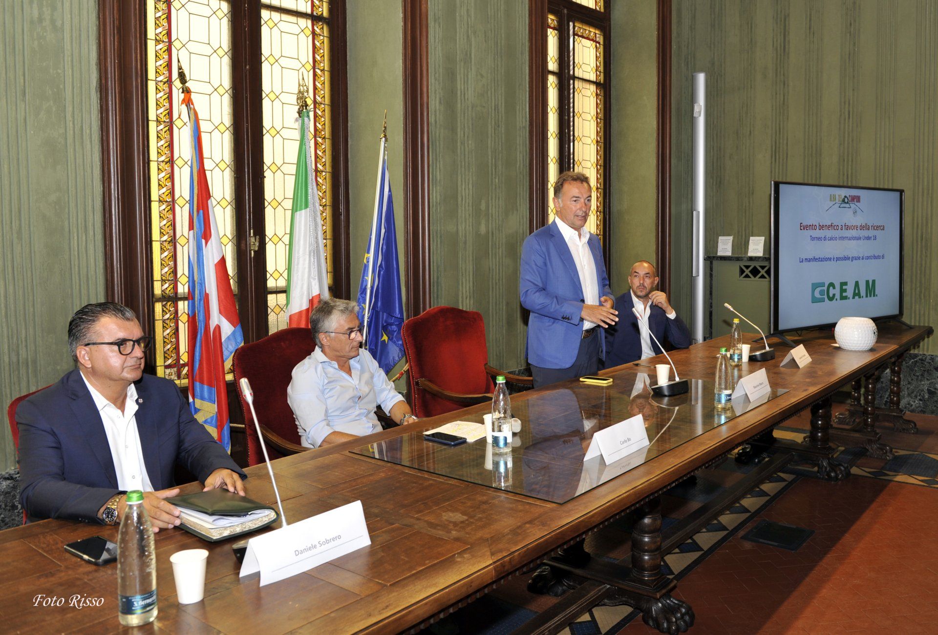 Un uomo parla seduto a un tavolo con altri, sullo sfondo delle bandiere. Uno schermo mostra del testo.