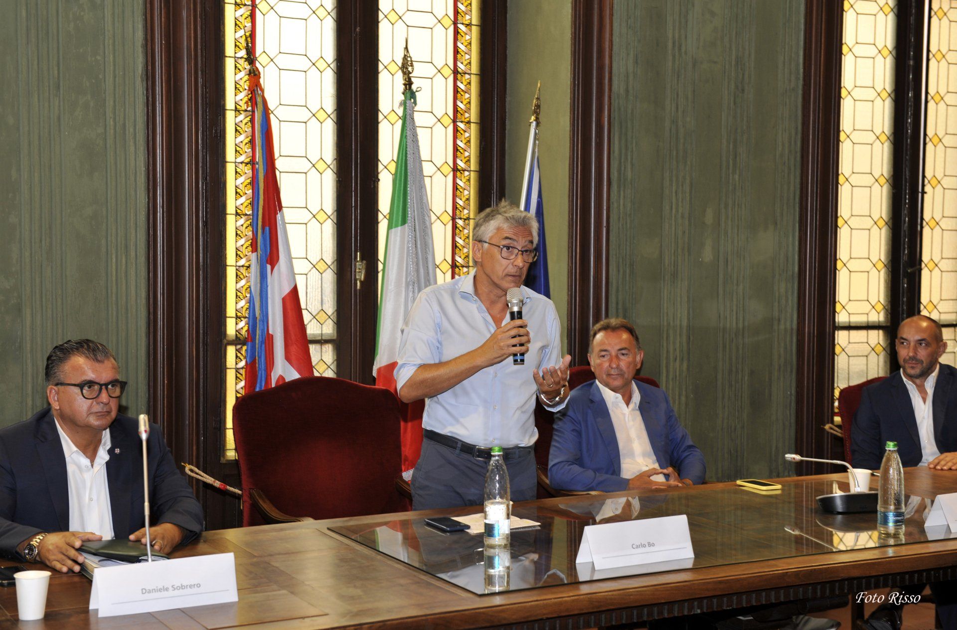 Uomo che parla a un tavolo con altri uomini; sullo sfondo bandiere e una stanza decorata.