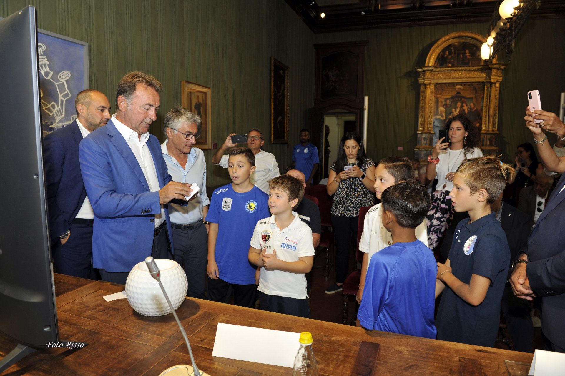 Un uomo in giacca blu firma autografi ai bambini in una stanza con dipinti e un grande monitor.