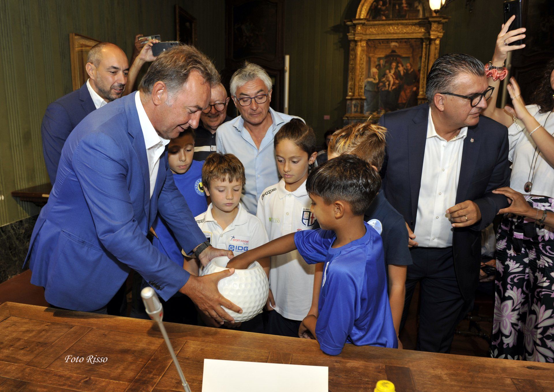 Uomini in giacca e cravatta, bambini in maglie blu, giocano con un pallone da calcio su un tavolo di legno. Sono presenti alcuni spettatori.