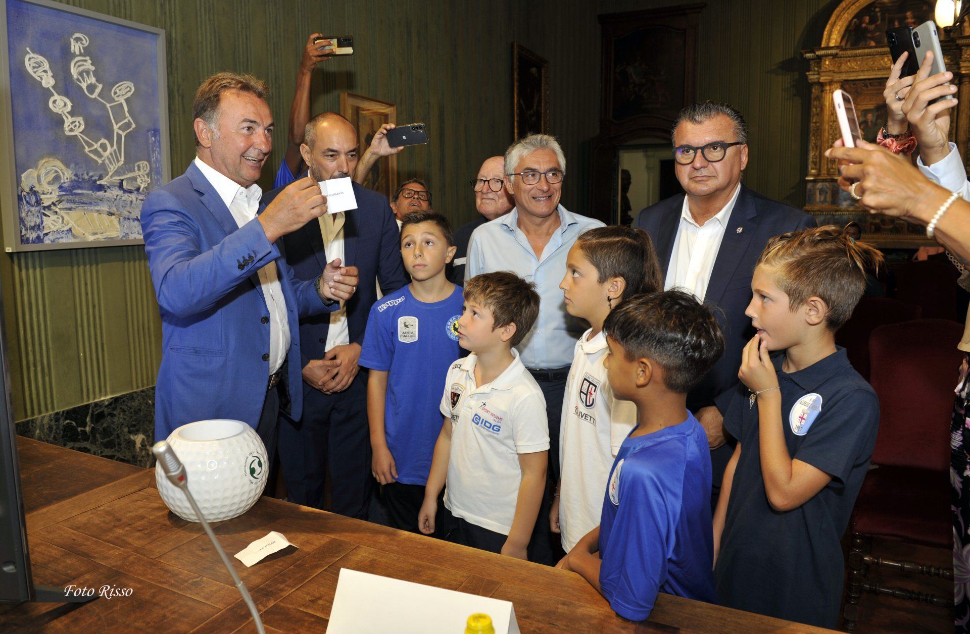 Un uomo in abito blu tiene in mano un foglio di carta sotto gli occhi dei bambini, all'interno di una stanza con un pallone da calcio decorativo.