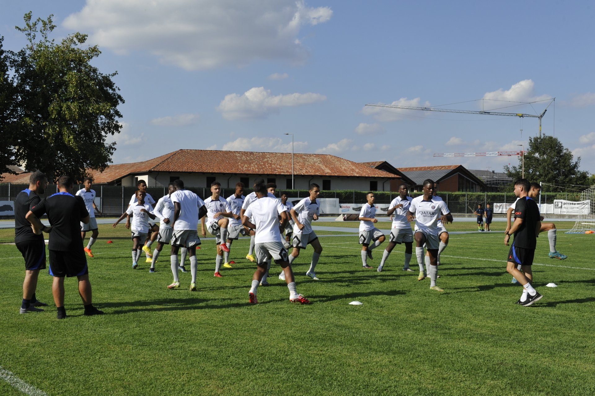La squadra di calcio in maglia bianca si riscalda su un campo verde, sotto gli occhi dello staff tecnico.