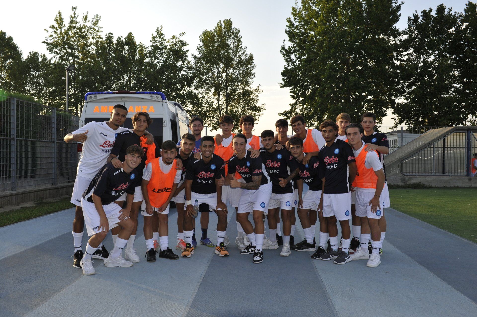 La squadra di calcio posa all'aperto su una pista blu, indossando maglie e pettorine arancioni, con alberi sullo sfondo.