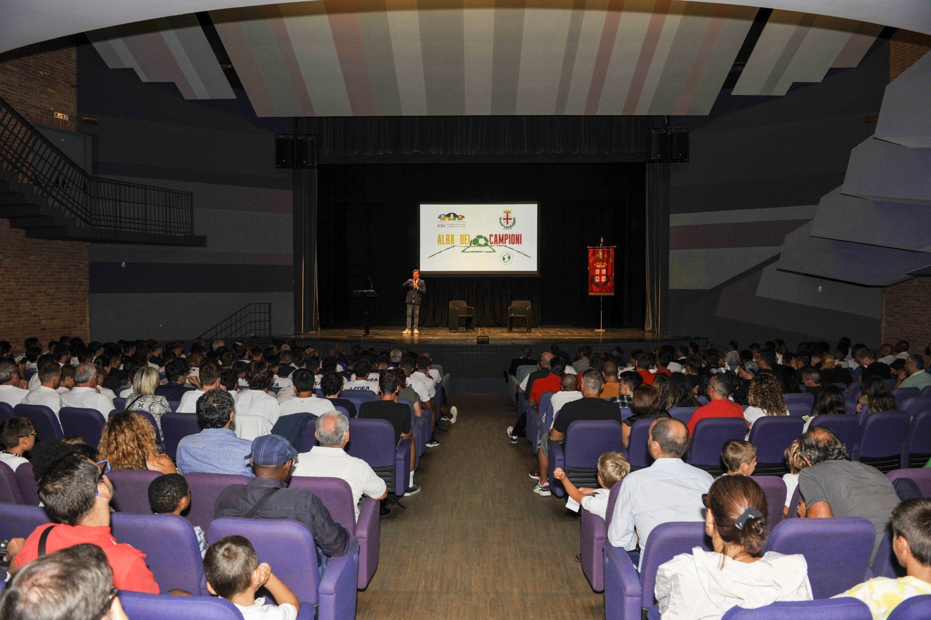 Il pubblico in un auditorium assiste a una presentazione su un palco; una persona sta parlando.