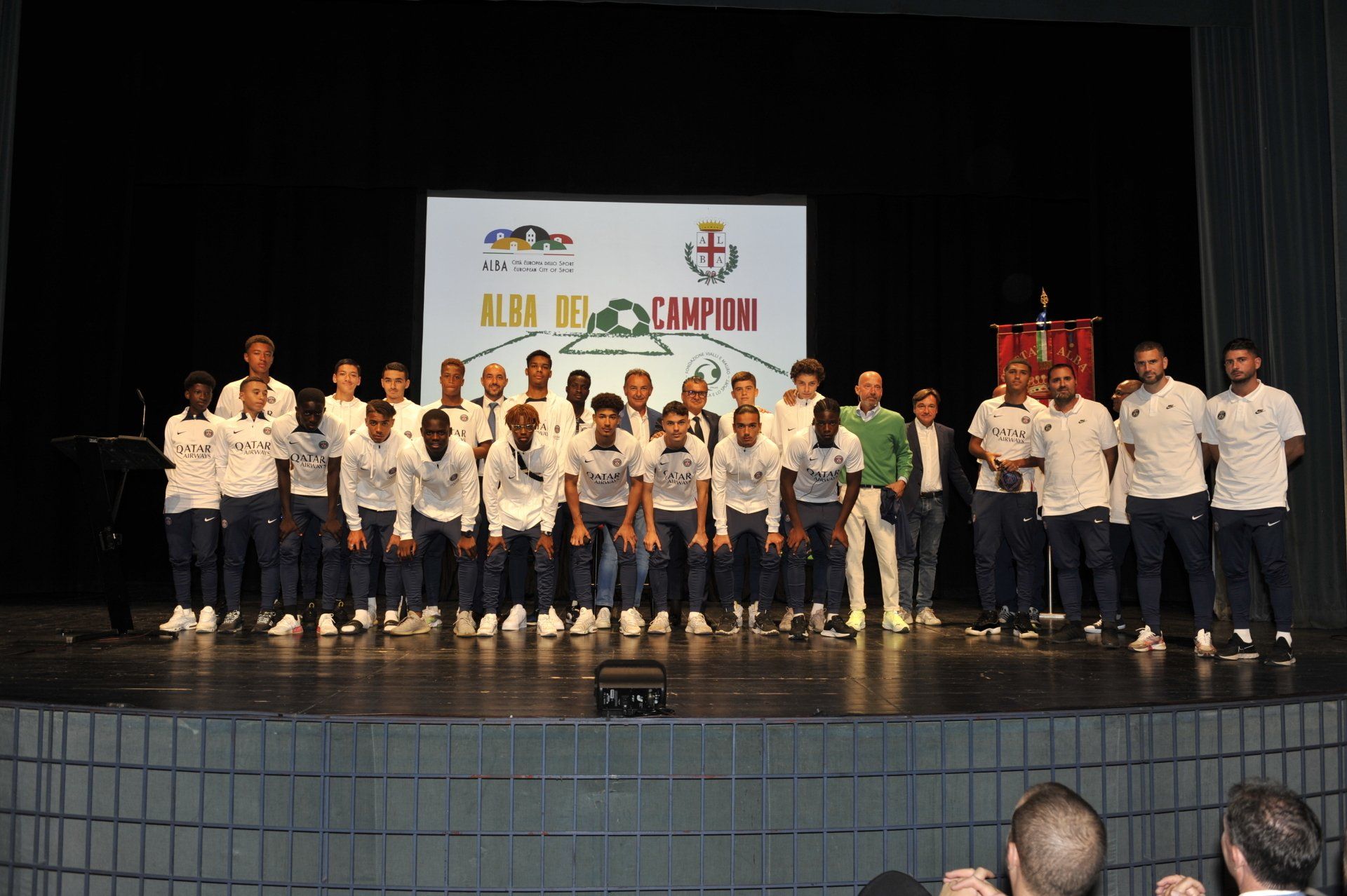 La squadra di calcio sul palco, con le divise bianche e blu, in piedi davanti a uno striscione.