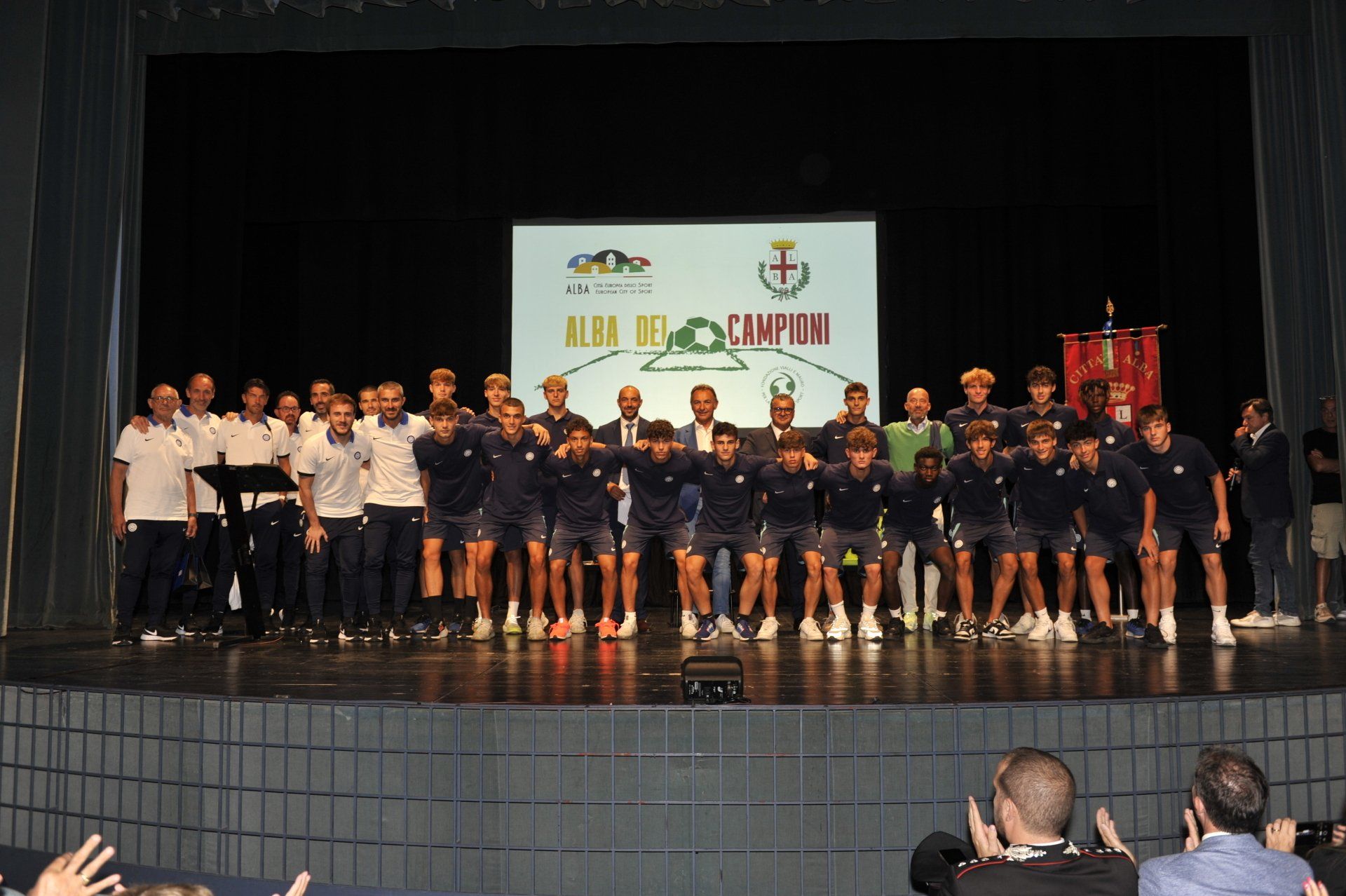 La squadra di calcio festeggia la vittoria sul palco, tenendo in mano il trofeo. Illuminata da uno schermo, la folla applaude.