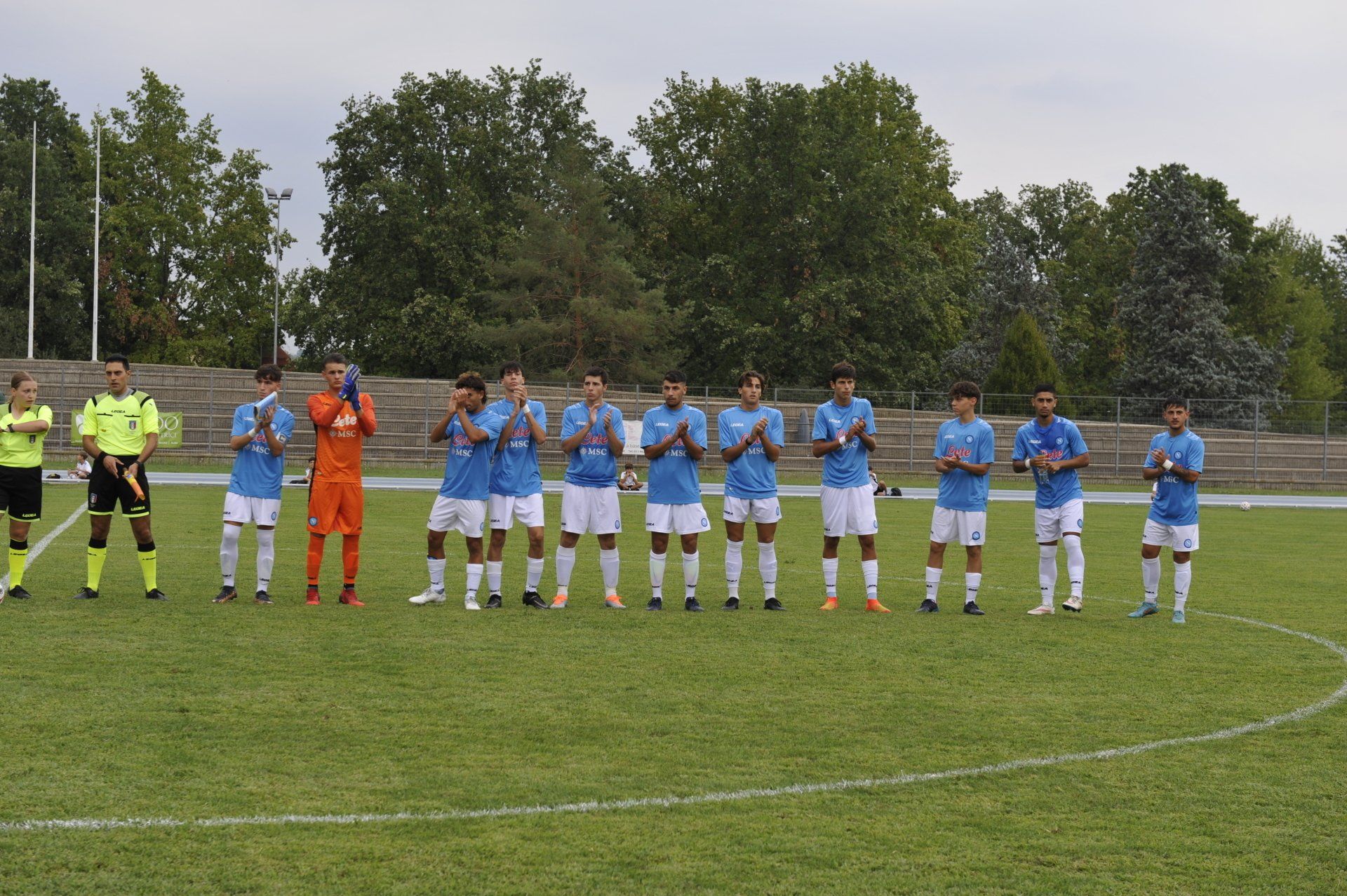La squadra di calcio in maglia azzurra e pantaloncini bianchi applaude su un campo verde. Gli arbitri sono in piedi nelle vicinanze.