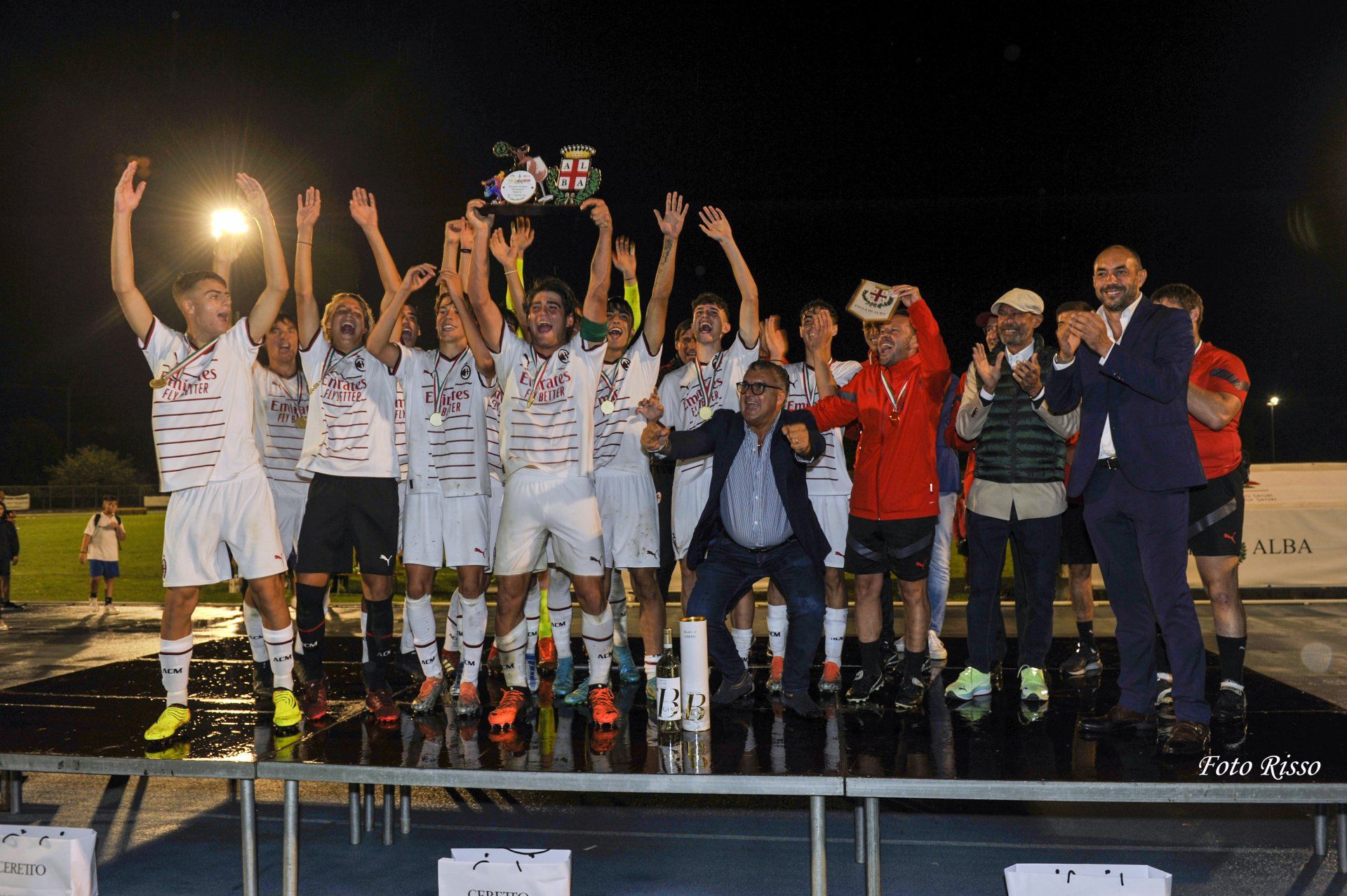Squadra di calcio che festeggia una vittoria, alzando un trofeo. Divise bianche, ambientazione notturna, tifo, palco.